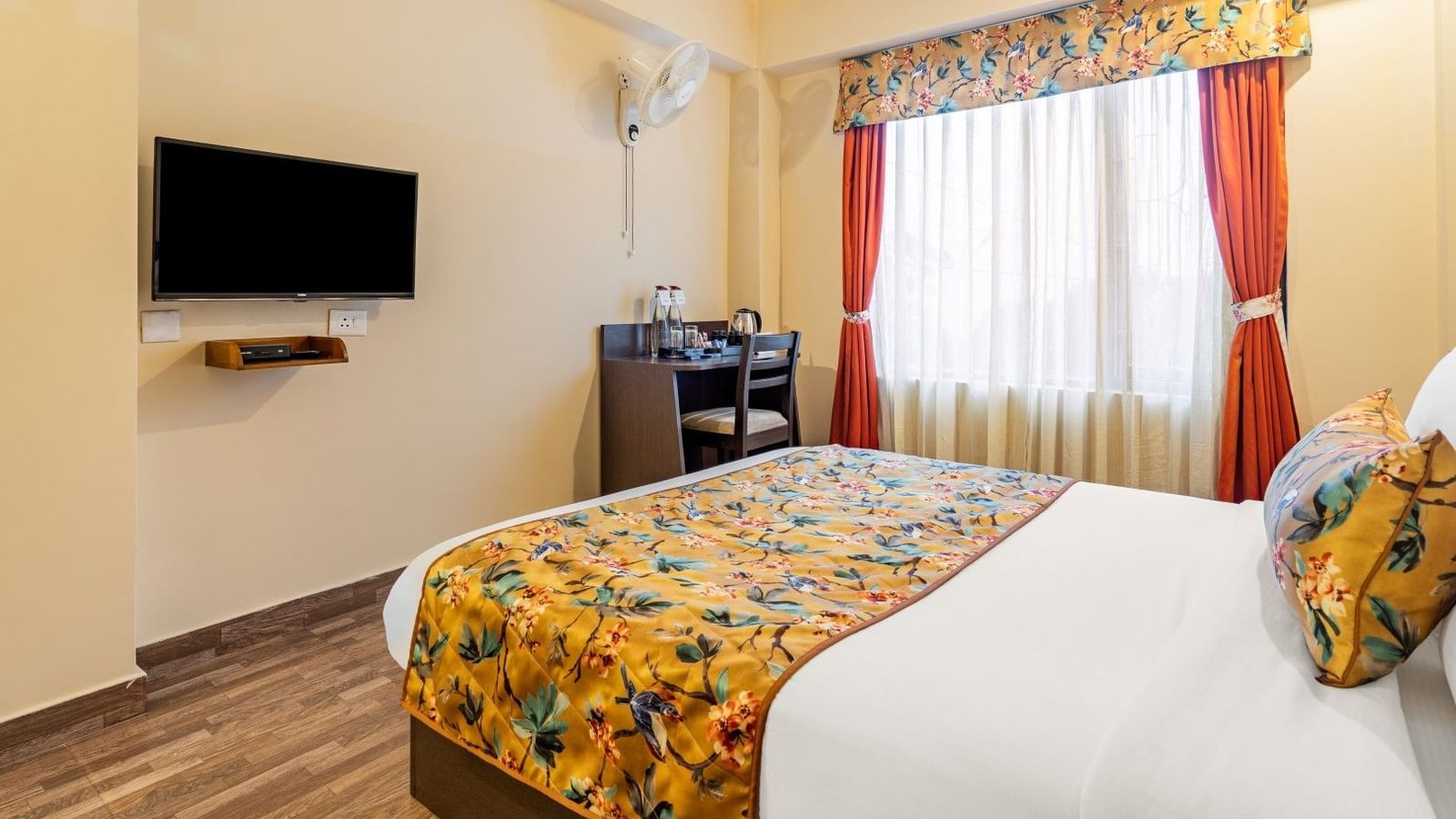 Cosy hotel room with a double bed, floral bedding, flat-screen TV, study desk with water bottles and kettle, and a large window with orange curtains - Summit Namnang Courtyard & Spa, Gangtok