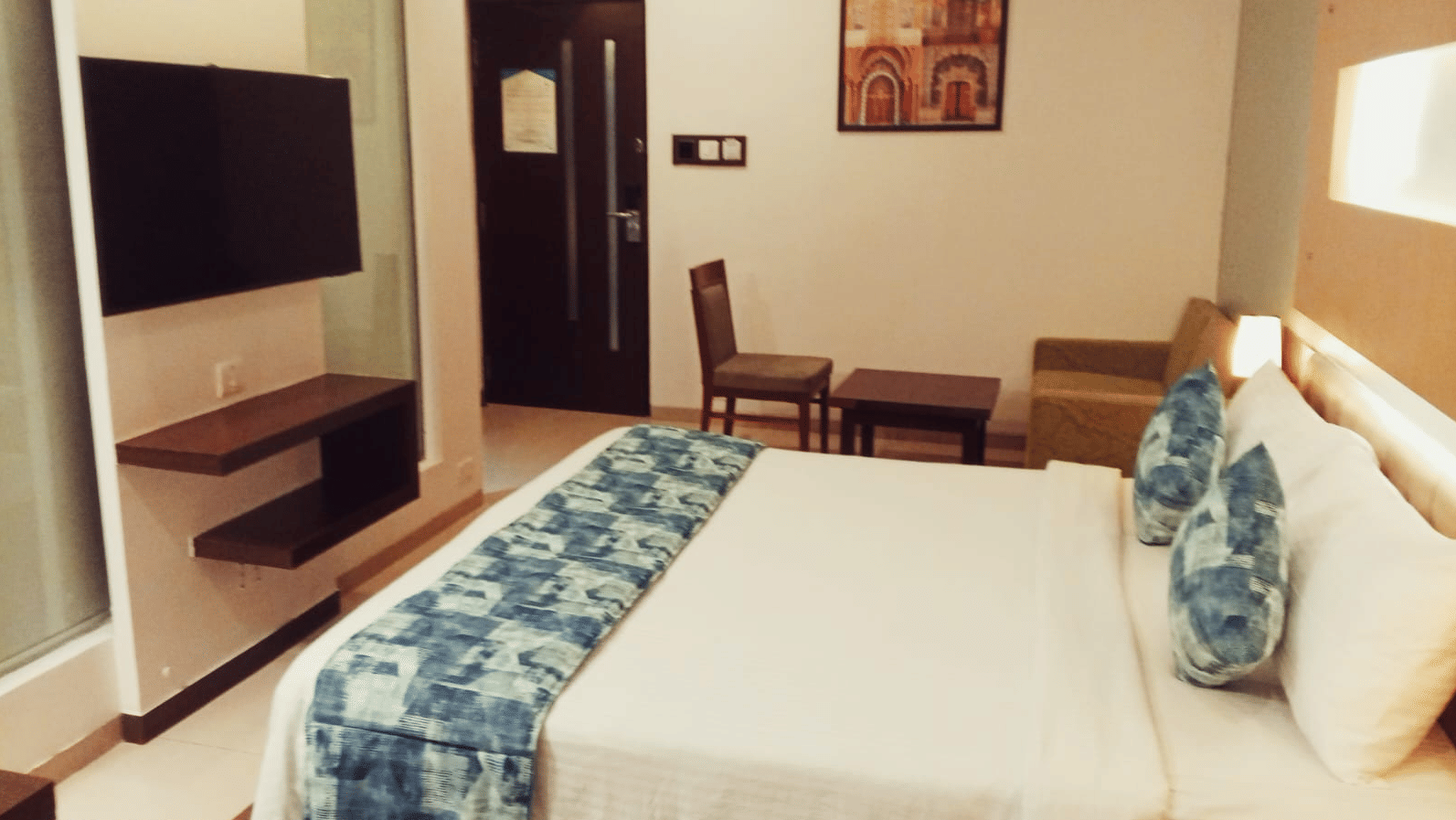 A hotel room layout showing a king size bed, a wall-mounted television, a wooden chair with a small side table, and an entry door at Hotel Mumbai House, Thane.