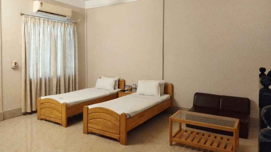 Interiors of a room at Lake Side Resort Neermahal featuring 2 wooden beds, an A/C unit, a small wooden table and tiled floors.
