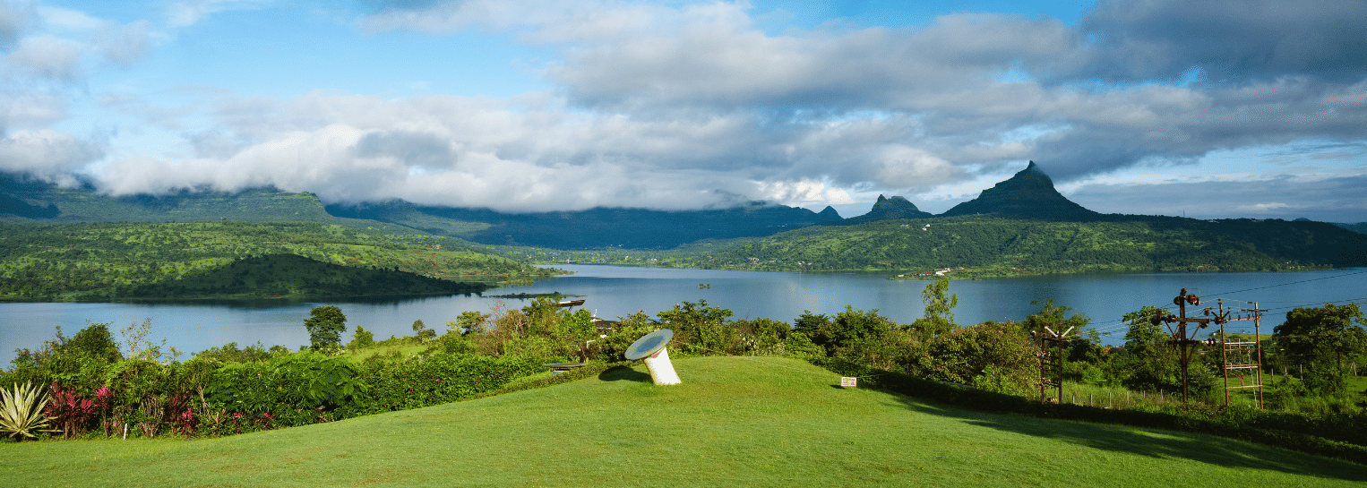 A wide panoramic landscape showing a calm blue lake, lush green lawn, and distant mountain peaks under a cloud-scattered sky.