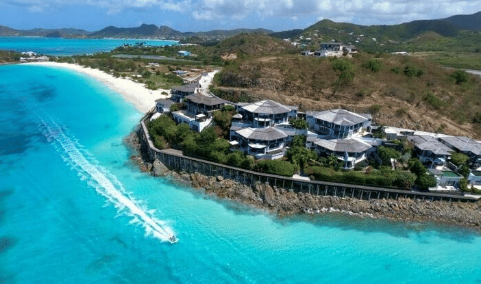 An aerial view shows a luxurious resort nestled along a pristine turquoise coastline with lush green hills.