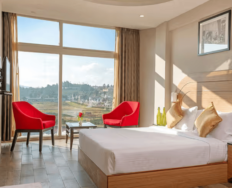 A hotel room with a large bed, two red armchairs, a side table, and a panoramic window overlooking a scenic landscape.