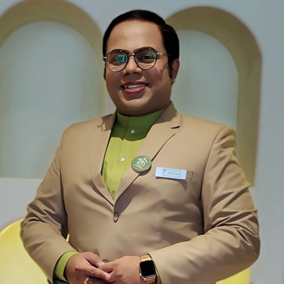 A man in a light brown suit jacket, green shirt, and blue name tag smiles with his hands clasped