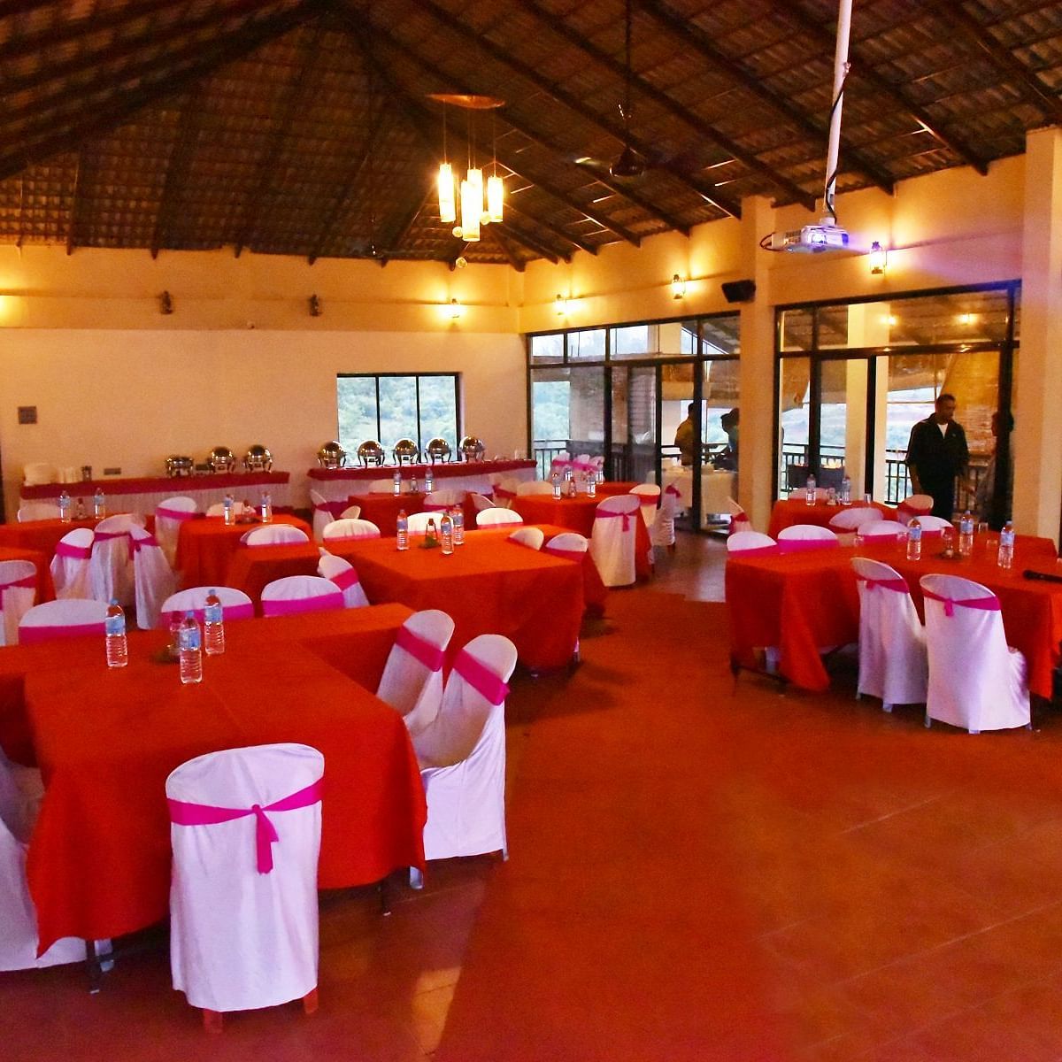 A banquet hall at Forest Escapes Koyna is prepared for a gathering with red tablecloths and white chair covers.