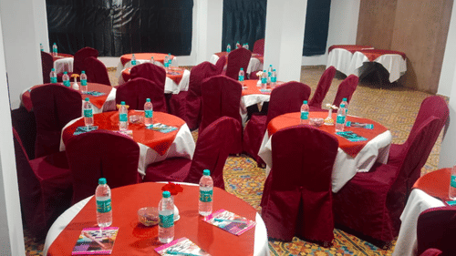 Event hall featuring round tables surrounded by chairs, each table having a water bottle, a book, a pen, and a flower vase
