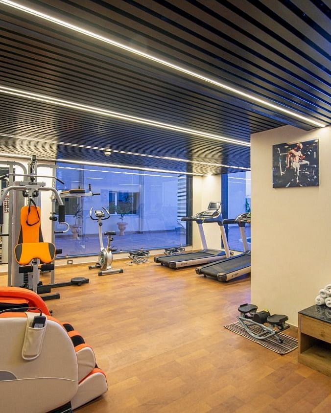 A modern gym area featuring various exercise equipment including treadmills, weight machines, a red and black punching bag, and a massage chair, with a wooden floor and a ceiling with strip lighting and a blue illuminated sign - Lords Inn Motikhavdi, Jamna