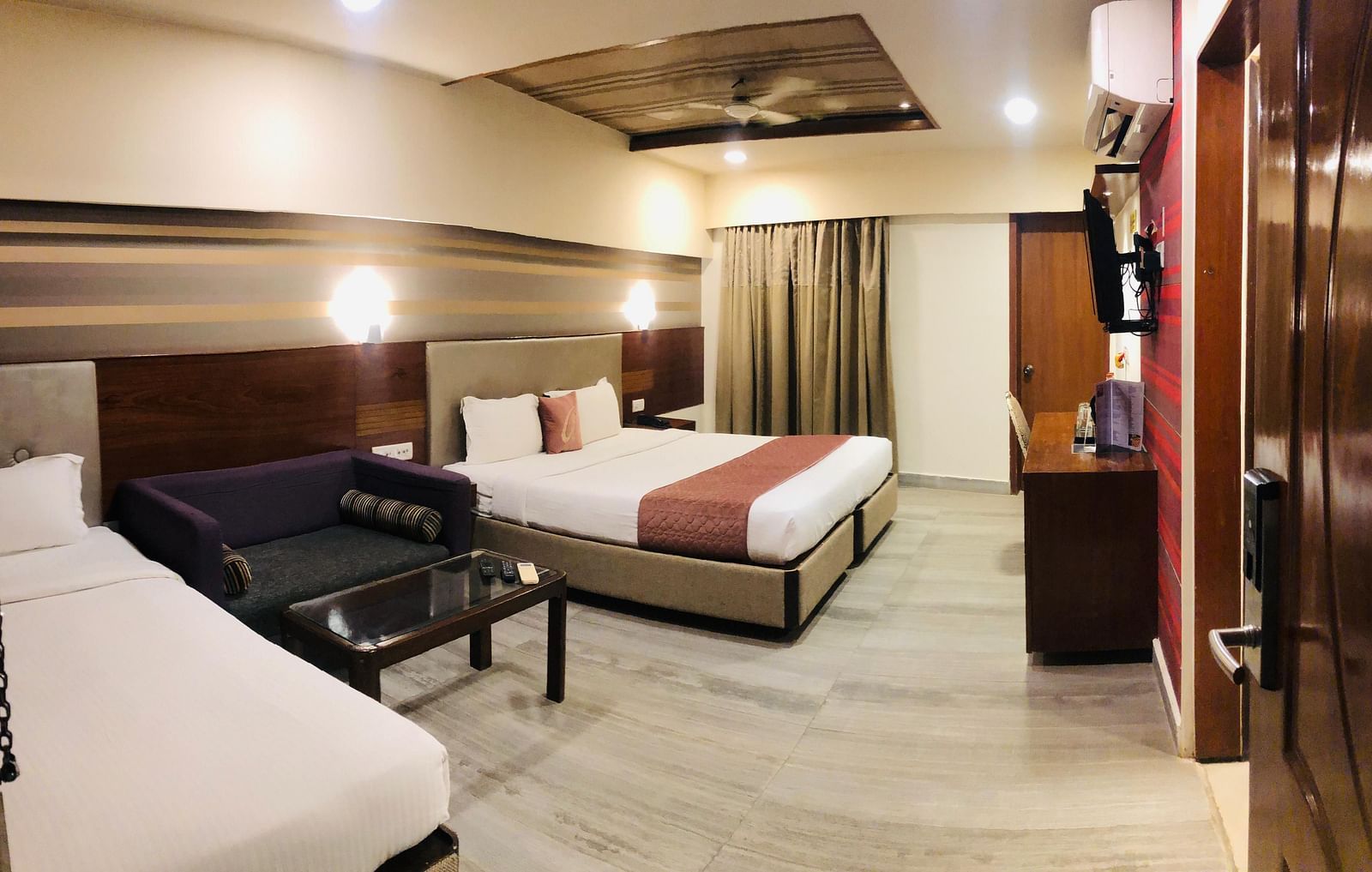 Hotel room interior showcasing a double bed, a dark coloured sofa, a glass-topped coffee table, and a work desk with a chair | Astra Hotels and Suites Kadubeesanahalli