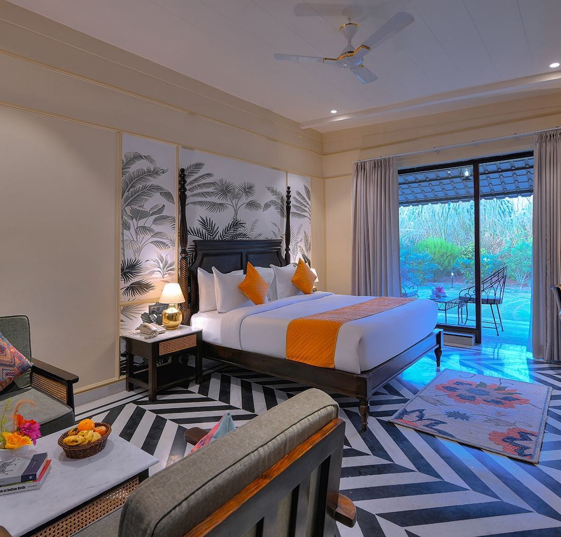 Superior Cottage featuring a four-poster bed with white linens, a colourful patterned runner and pillows, a patterned armchair, a wall-mounted TV, and sliding glass doors leading to a balcony - Singhvilas, Ranthambore