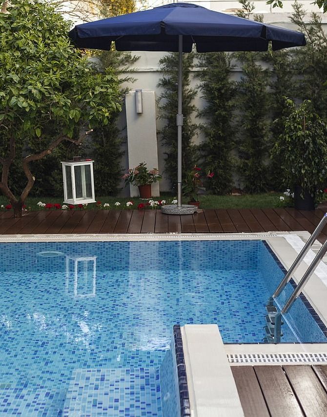 A swimming pool with a wooden desk around it with a tall standing umbrella on the deck.