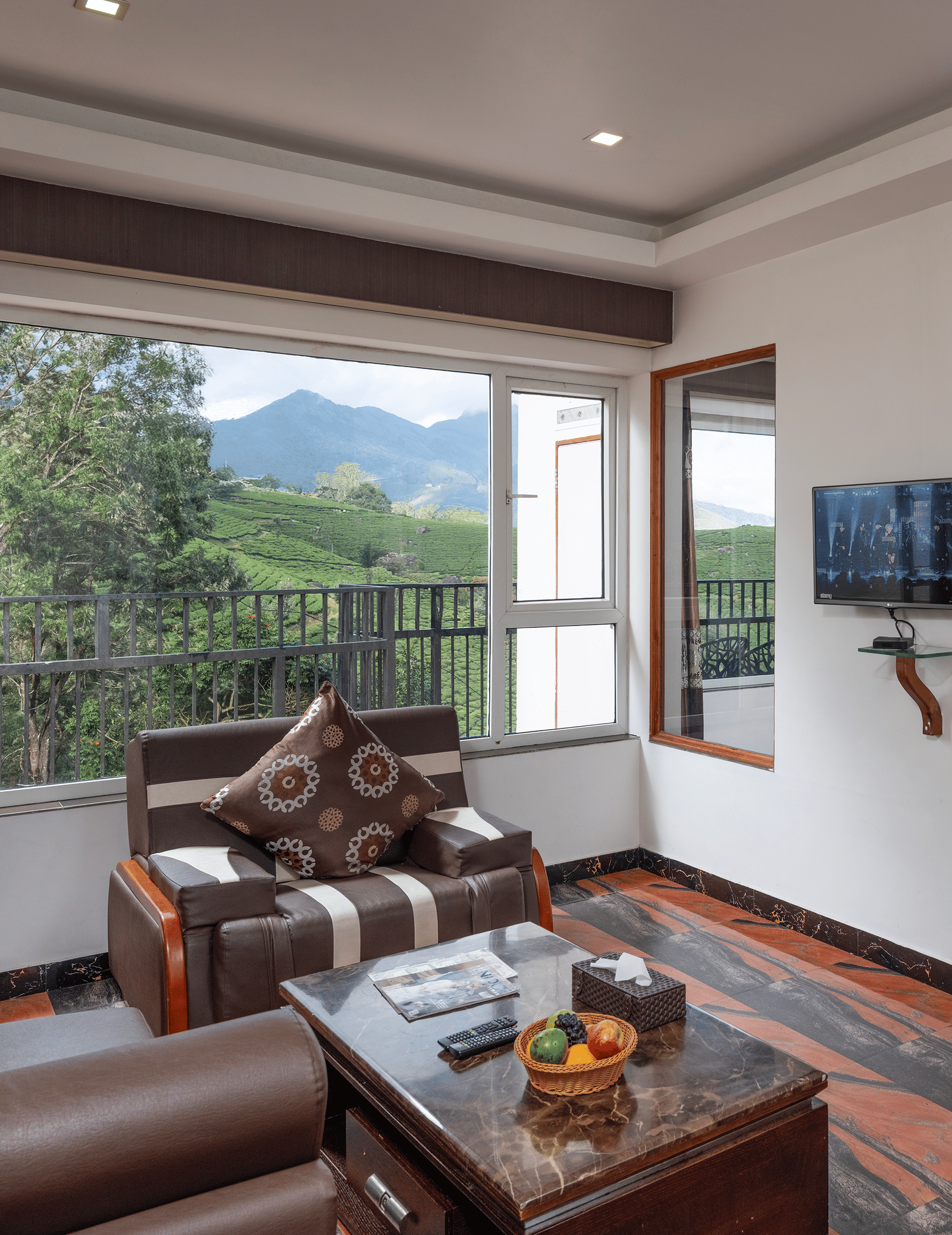 The spacious interior of the Premium Room at Parakkat Nature Hotel and Resorts, Munnar featuring a separate living area with wooden furnishing, an opening towards bedroom, and large glass windows overlooking the greenery.