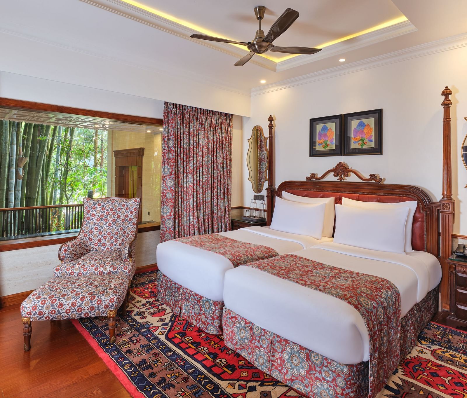 Royal Villa featuring a wooden bed with white bedding, a patterned armchair, a decorative rug, a ceiling fan, framed pictures, and a window with bamboo trees, complemented by a patterned curtain