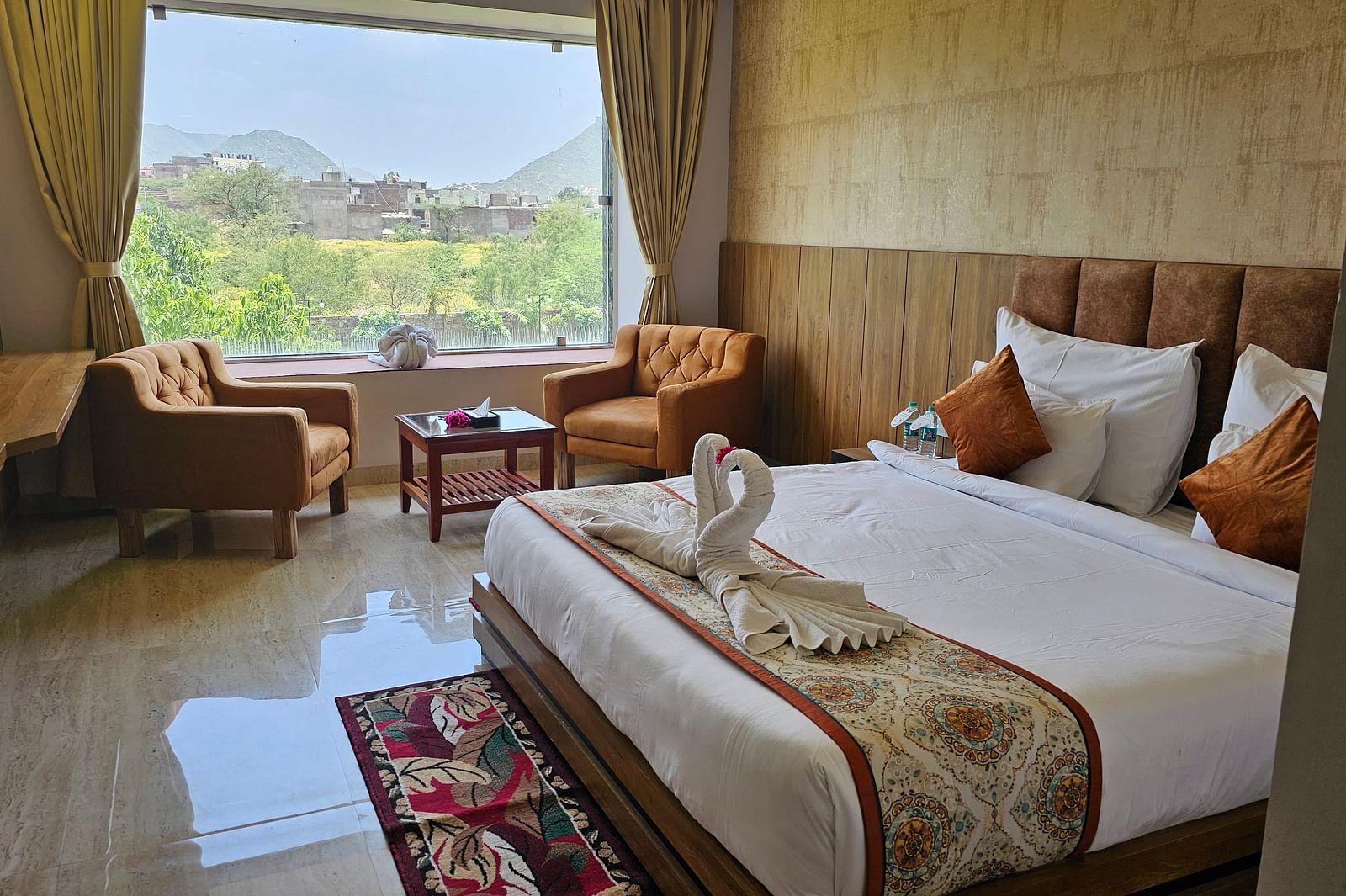 A bright and spacious resort room at Bamboo Saa Mulberry Resort, Pushkar, featuring a large window with a view of the outdoors including a made bed with a patterned runner, two armchairs, and a small table.