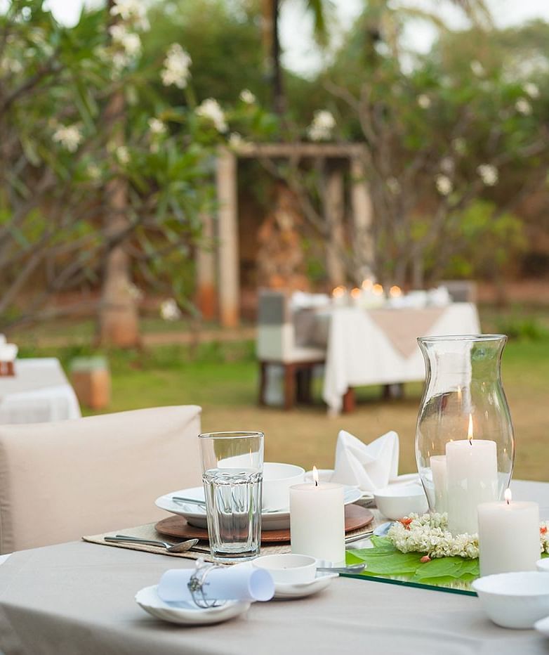 An outdoor dining setup with white tablecloths and chairs, green lawn and trees in the background - Shreyas Retreat