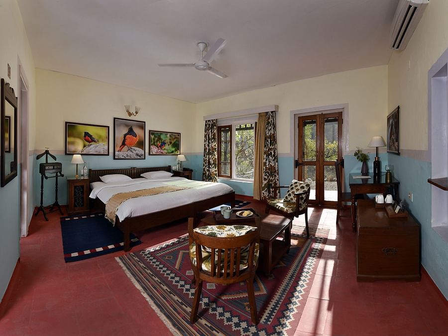 A spacious and elegantly decorated room featuring a large bed, vintage wooden furniture, and vibrant artwork on the walls. Sunlight streams through the windows, highlighting the cosy seating area and colourful rugs.