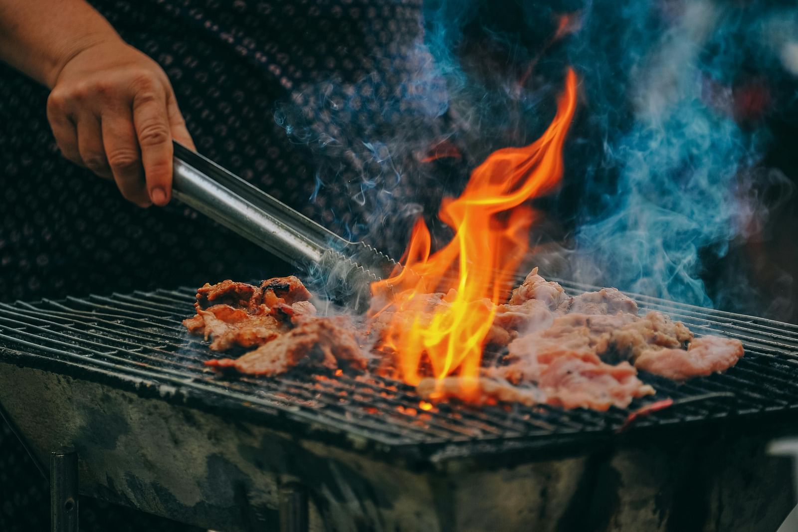 A hand holding tongs over a grill with meat, with flames and smoke rising.