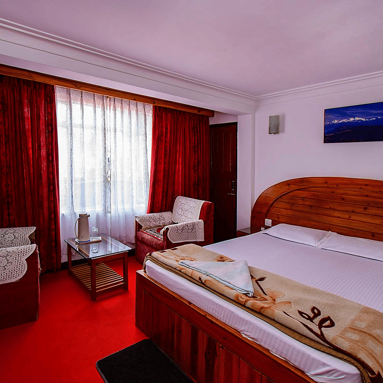 Hotel bedroom featuring a double bed, red carpet, white walls, and a small sitting area with patterned chairs at Hotel Dolphin Darjeeling.