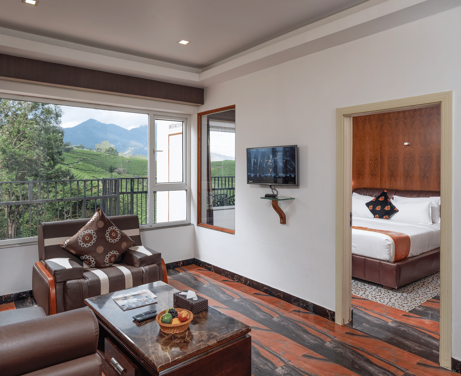 The spacious interior of the Premium Room at Parakkat Nature Hotel and Resorts, Munnar featuring a separate living area with wooden furnishing, an opening towards bedroom, and large glass windows overlooking the greenery.