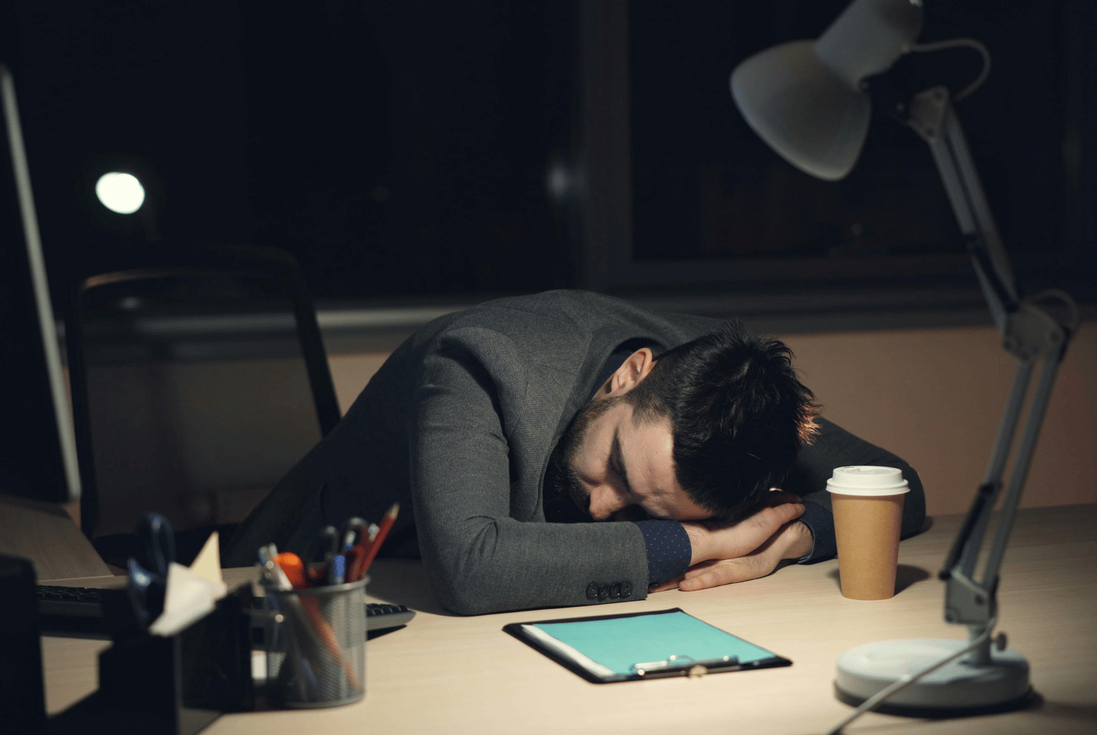 A person lying heads down on a table that has a notepad, penstands, coffee mug, and a table lamp, placed on it.