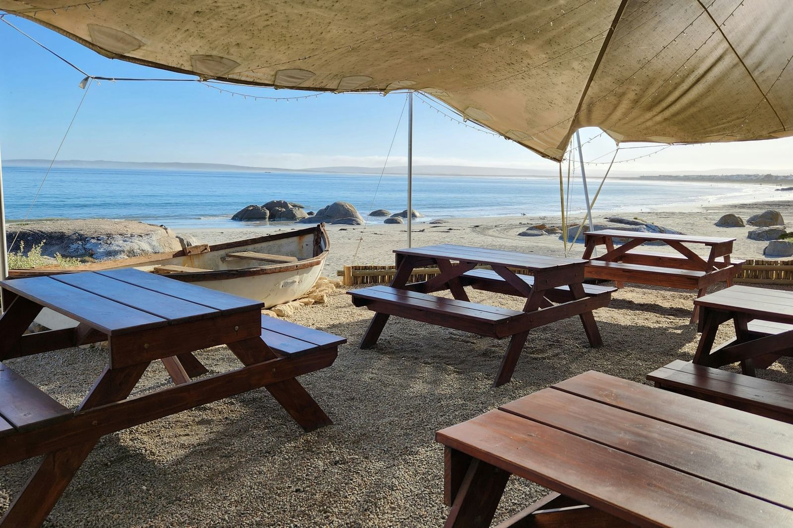 A view of a beach-side restaurant with a canopy above and wooden tables below it.