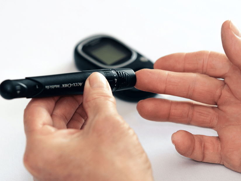 Hands using a lancing device on a fingertip, with a blood glucose meter in the background