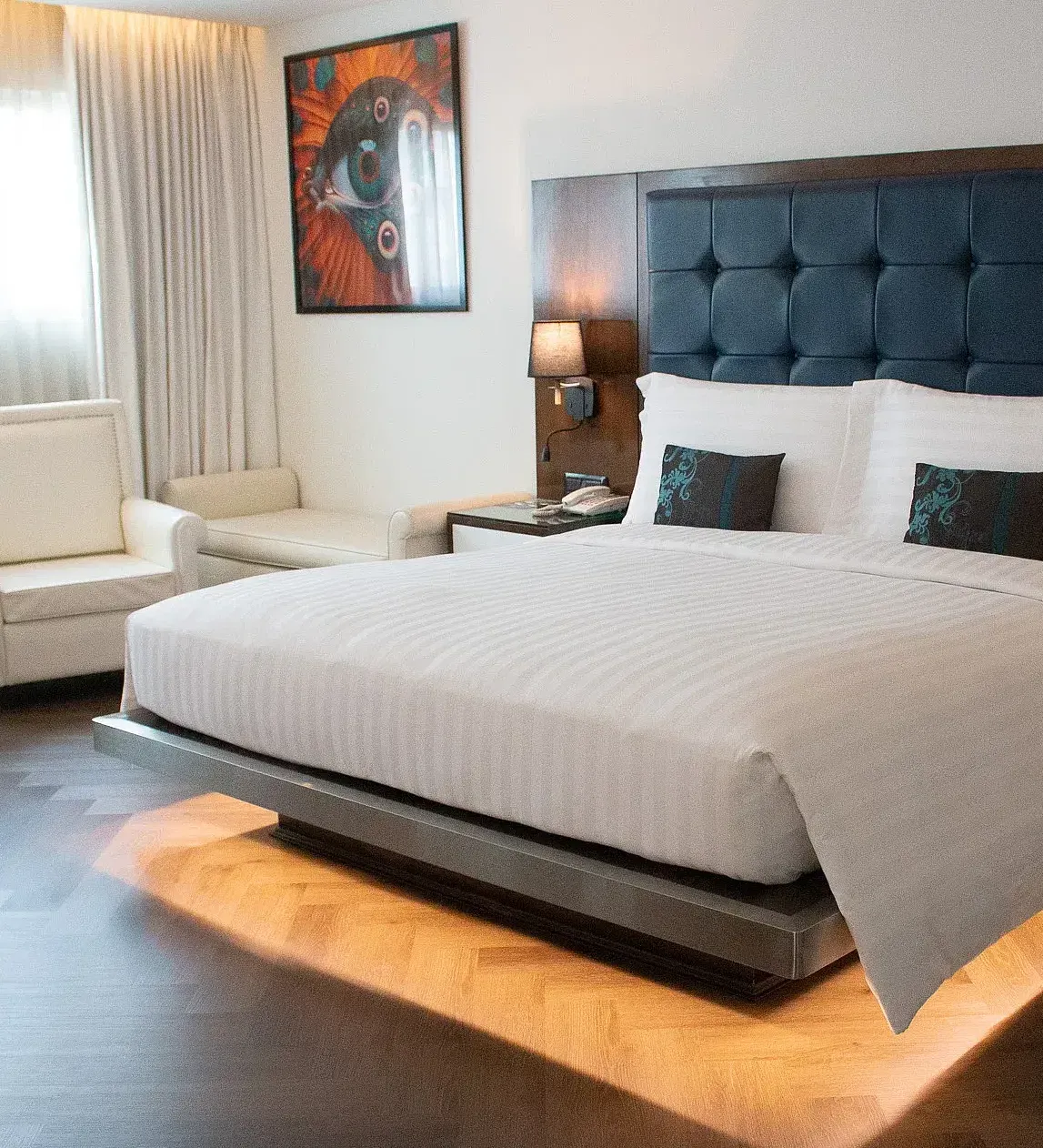 Luxurious hotel suite bedroom with illuminated platform bed, tufted headboard, vibrant artwork, cream armchairs, and warm ambient lighting throughout at Night Hotel Bangkok.
