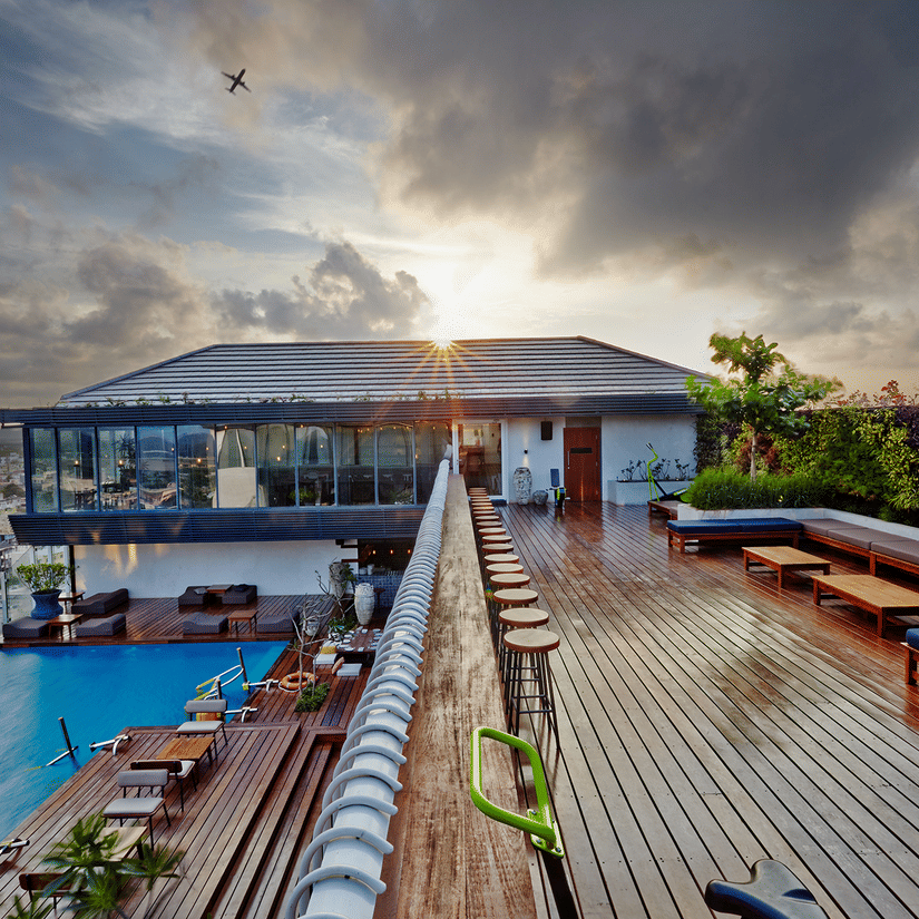 Aqua gym featuring a wooden deck, infinity pool, seating areas with city views in front under cloudy sky at sunset at Hablis Hotel, Chennai