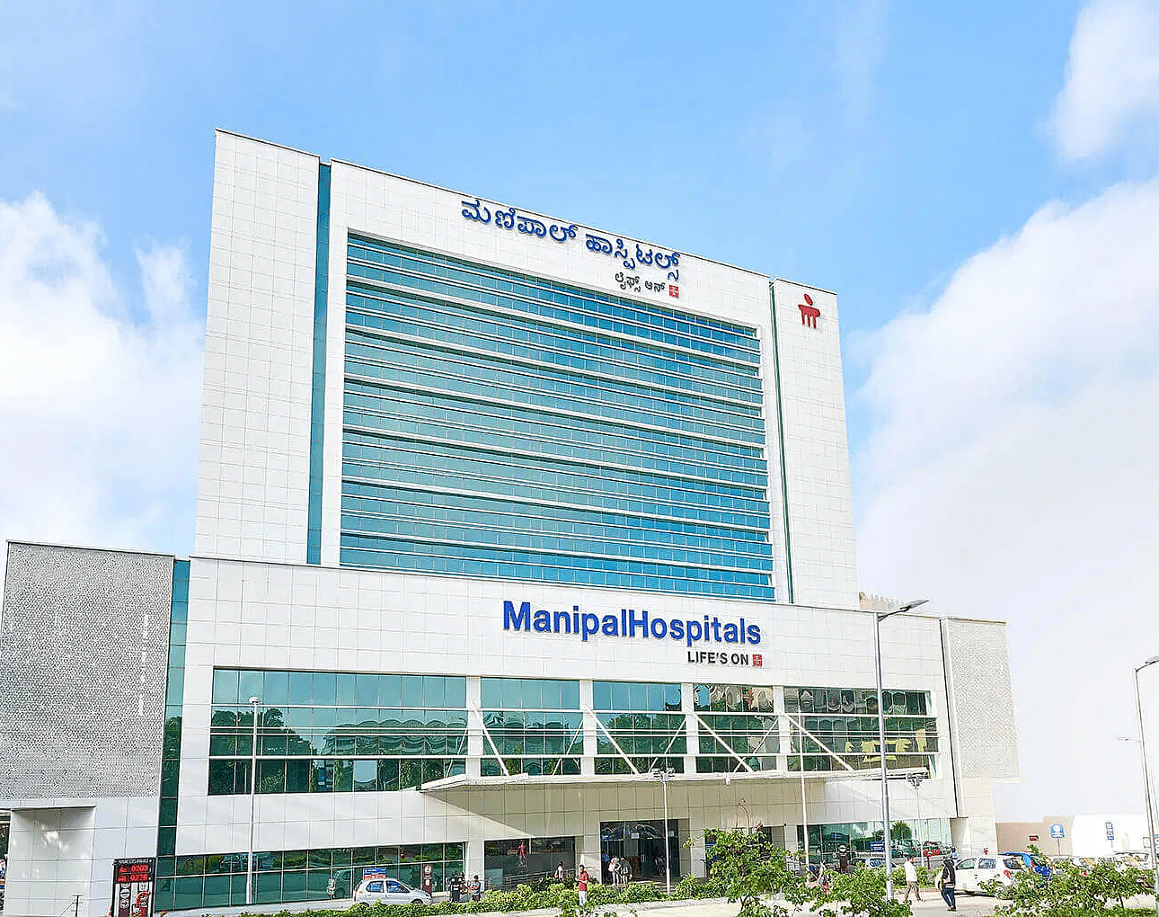 A modern, multi-storey hospital building with the Manipal Hospital's sign and surrounding green trees under a blue sky.