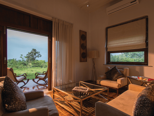 Sun illuminating the interiors of a room at The Serai Bandipur featuring a sofa set and a patio seen through the doorway with a couple of chairs facing the trees outside