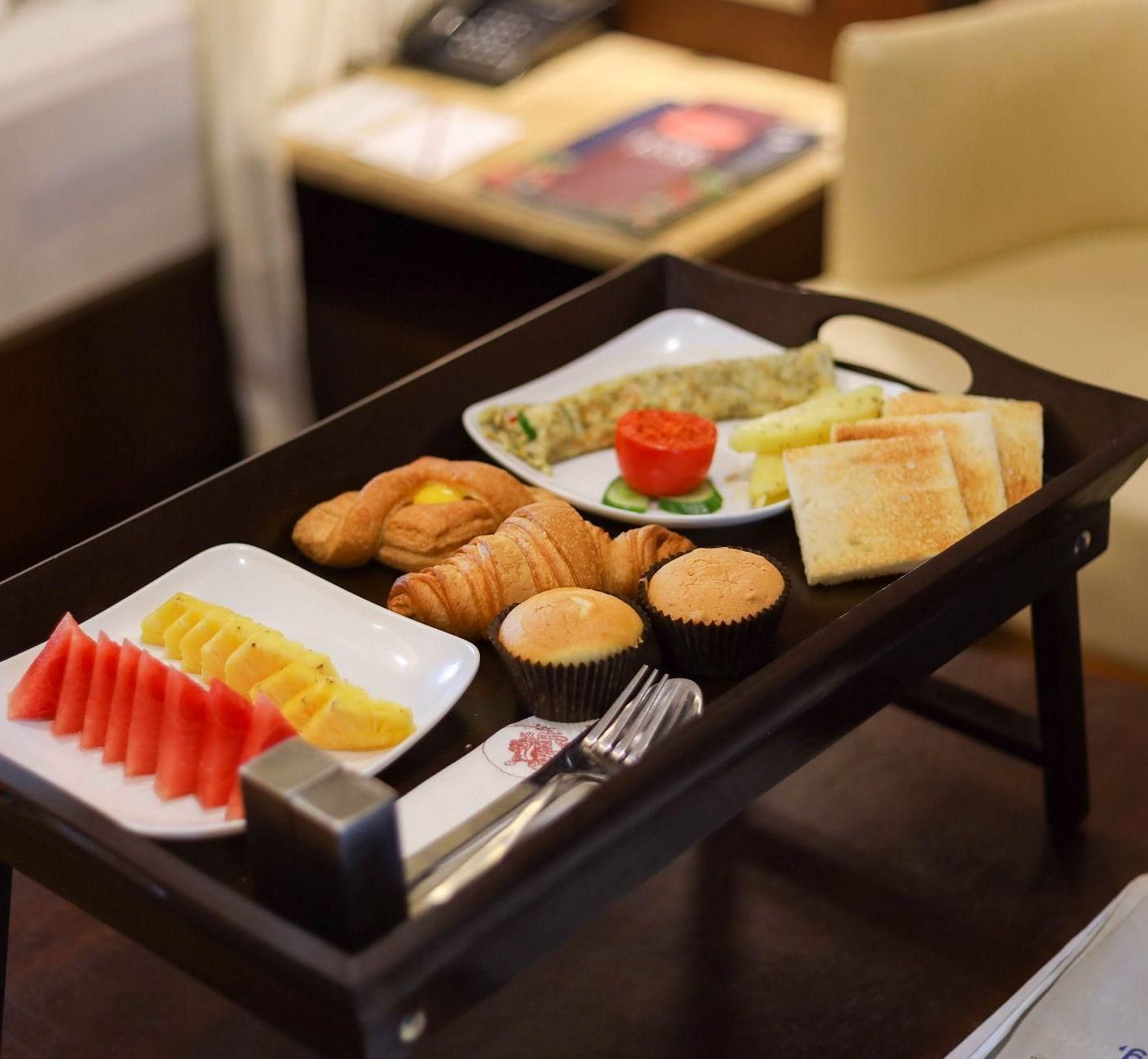 Room service breakfast tray with toast, muffins, croissants, omelette, fresh fruit, and cutlery set, placed on a table iin Avana Resort