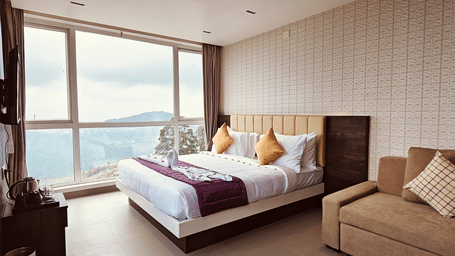 A bedroom featuring a double bed, a brown sofa, and a large window overlooking hills at Bliss Casa Mila By Bergamont Hotels, Kodaikanal.