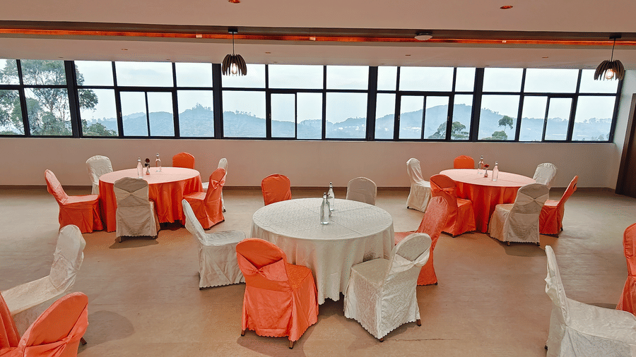 Multiple round tables with peach covers are placed near large windows showing hills at Bliss Casa Mila By Bergamont Hotels, Kodaikanal.