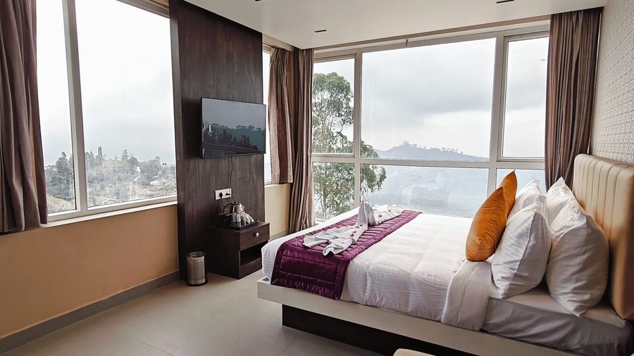 A bedroom features a television on a dark panel and large windows showing trees and hills at Bliss Casa Mila By Bergamont Hotels, Kodaikanal.