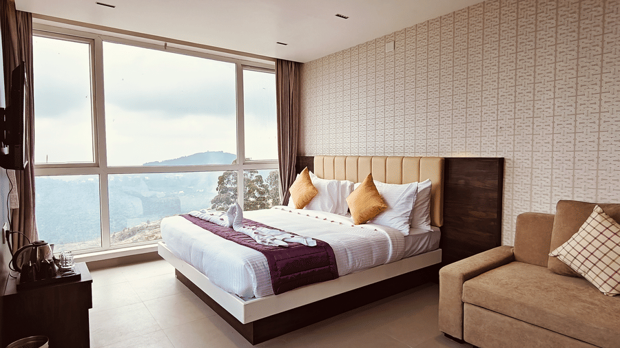 A bedroom featuring a double bed, a brown sofa, and a large window overlooking hills at Bliss Casa Mila By Bergamont Hotels, Kodaikanal.