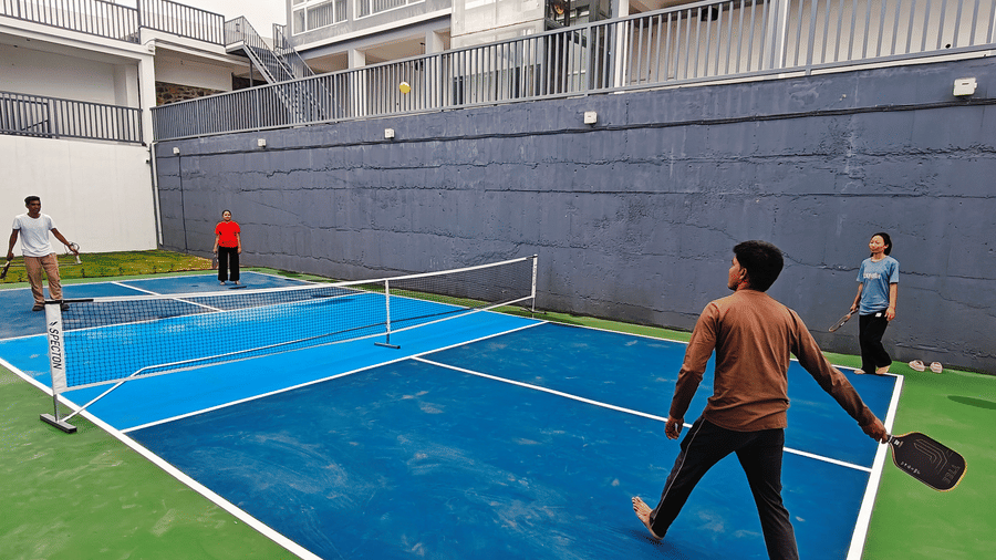 4 people playing a game of pickleball on a blue outdoor court with a grey wall behind at Bliss Casa Mila By Bergamont Hotels, Kodaikanal.