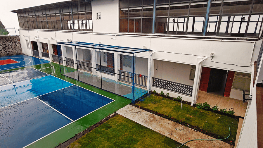 Elevated view of 2 blue and green outdoor sports courts with a white building and grass sections at Bliss Casa Mila By Bergamont Hotels, Kodaikanal.