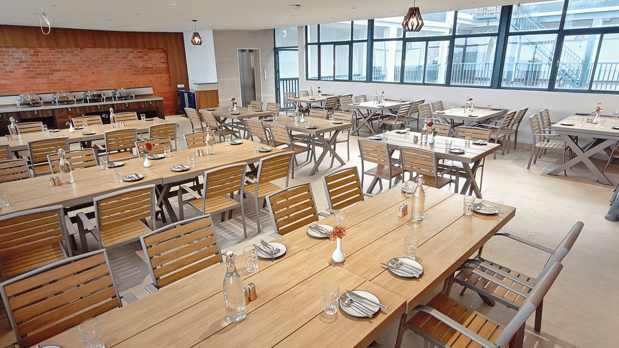 A restaurant area contains several rows of wooden tables and chairs with glass windows at Bliss Casa Mila By Bergamont Hotels, Kodaikanal.