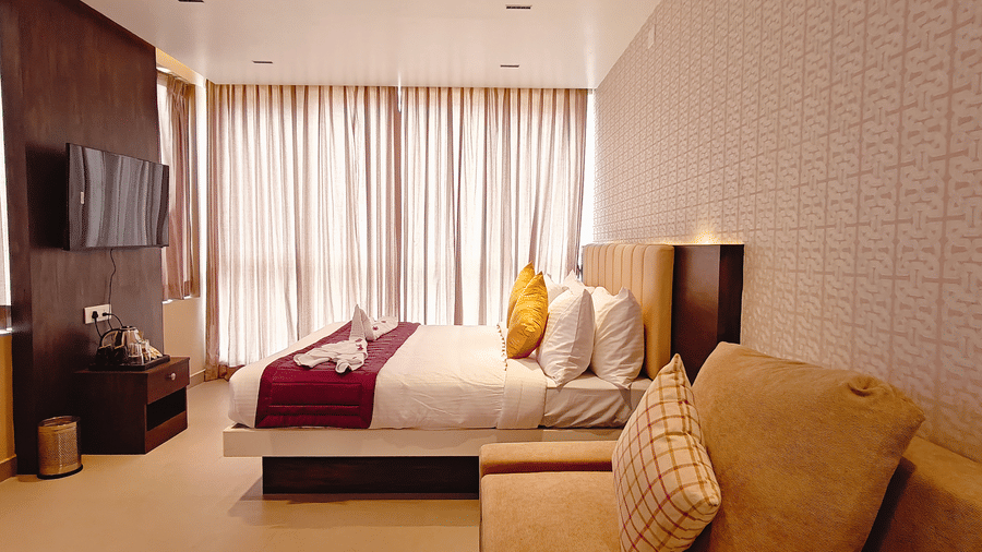 A room contains a white bed with purple accents, a beige sofa, and a television mounted on a dark wooden wall at Bliss Casa Mila By Bergamont Hotels, Kodaikanal.