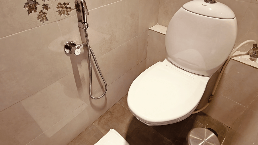 A white toilet and a hand-held shower head against beige tiled walls at Bliss Casa Mila By Bergamont Hotels, Kodaikanal.