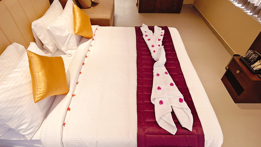 A white bed features towel art decorated with pink petals, placed over a purple bed runner with 2 yellow cushions at Bliss Casa Mila By Bergamont Hotels, Kodaikanal.