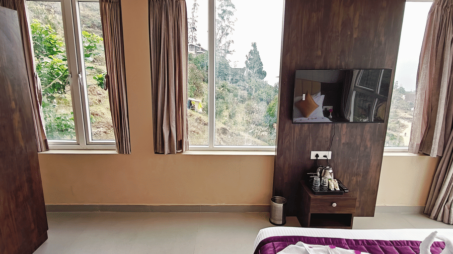 A dark wooden wardrobe stands next to 3 windows draped with light brown curtains showing an outdoor view at Bliss Casa Mila By Bergamont Hotels, Kodaikanal.