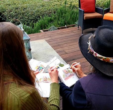 Two women sitting outdoors with a scenic ocean view, engaged in painting or sketching, with one wearing a black hat adorned with flowers.