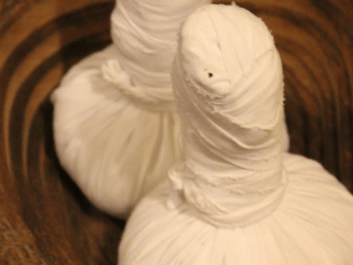 Two herbal compress balls (potlis) rest in a dark wooden bowl.