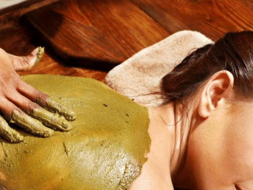 A woman lies face down receiving a full-back body wrap, with a therapist applying a thick herbal paste.