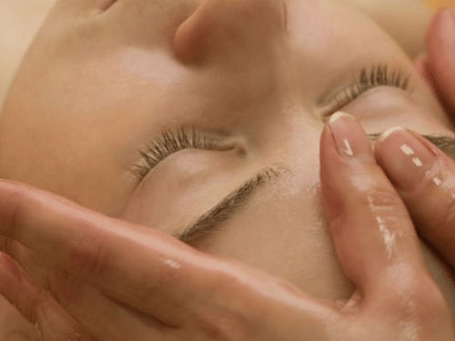 A person with their eyes closed is receiving a Ayurvedic facial massage around the temples and eyes with oil.