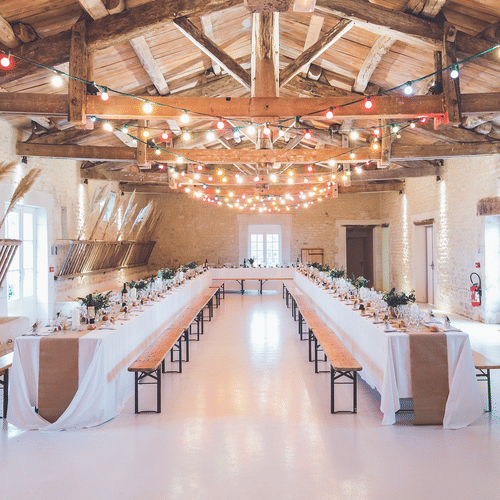  The image shows a banquet hall with a rustic aesthetic, featuring exposed wooden beams, stone walls and string lights.