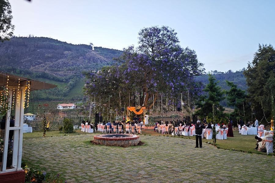 Wide shot of an outdoor wedding venue with guests seated around a central stage under a large tree with purple flowers in Coonoor - Ibex Stays & Trails, Coonoor (Leewood).
