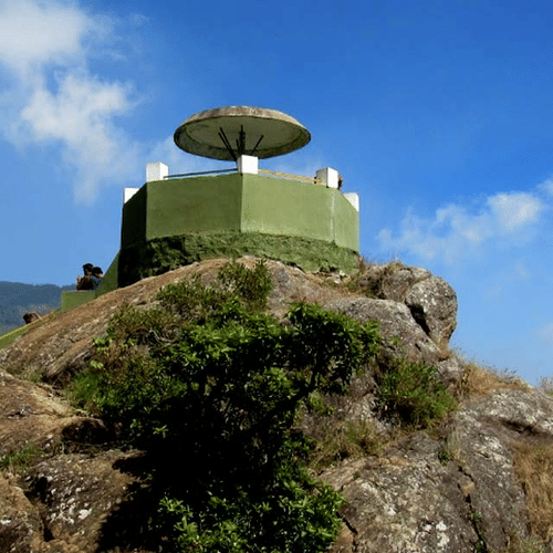 A stone structure with a circular platform perched on top of a rocky hill, set against a bright blue sky with scattered clouds.
