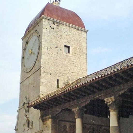 A historic clock tower with a red-roofed top and stone walls, part of an old building under a blue sky.