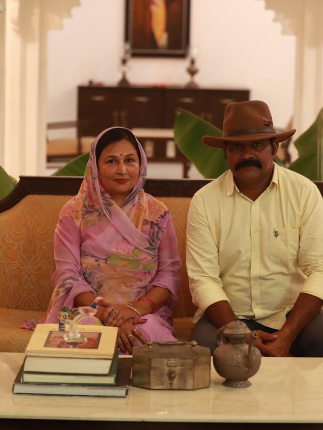 A man in a brown hat and a woman in a pink saree are sitting on a tan sofa in a traditional room.