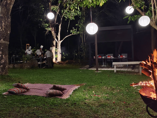 An outdoor bonfire in a metal pit on the right. A blanket sits on the grass, surrounded by trees and hanging globe lights.